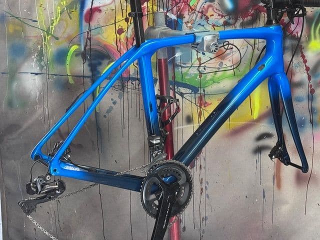 Fully built bike with a frame refinished in gloss blue, photographed against the shop's paint-splattered backdrop.