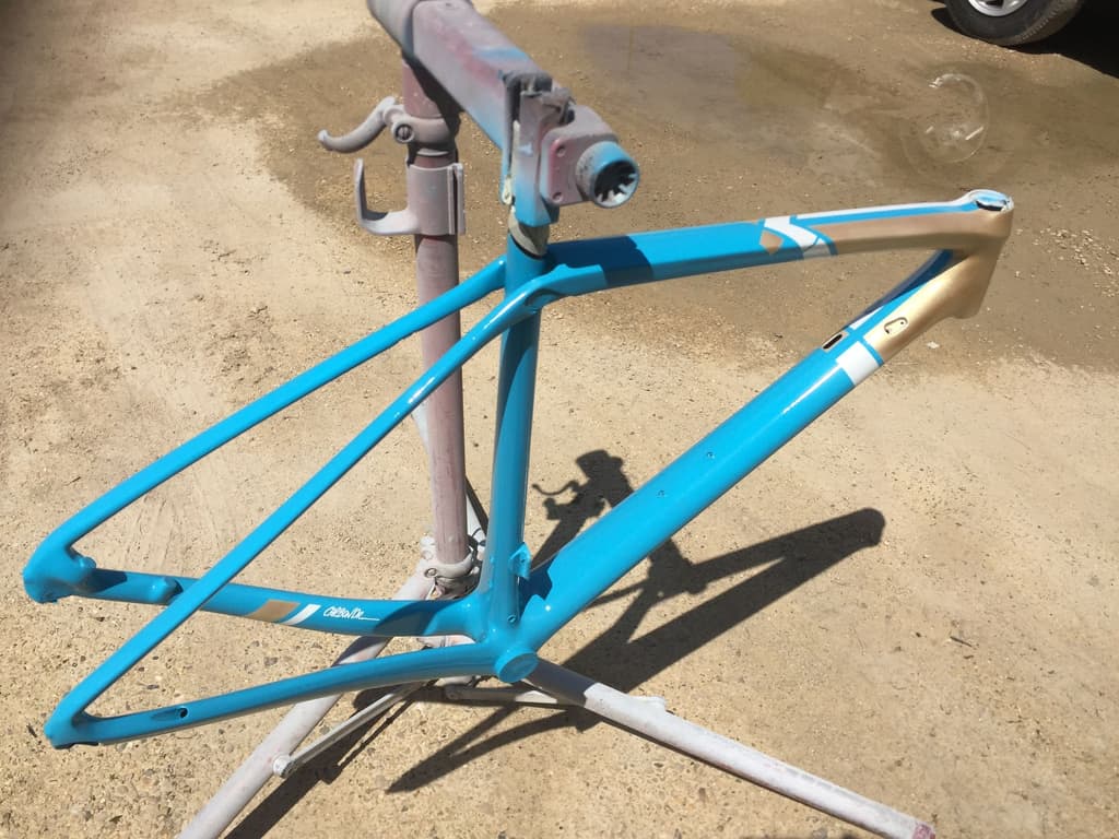 Frame refinished in a bright cyan/turquoise with a thin gold pinstripe along the top tube, photographed outside on a driveway.